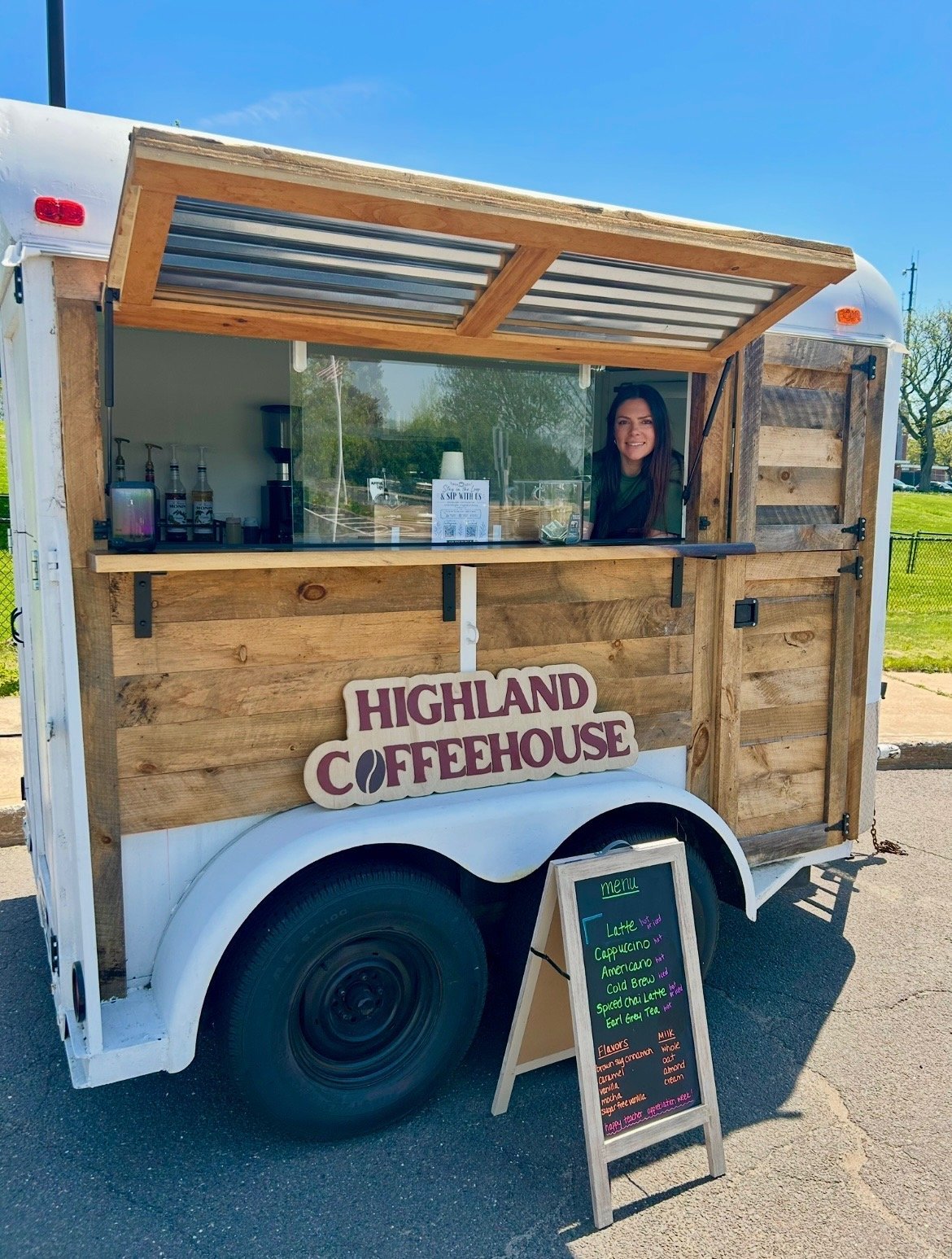 Mobile Highland Coffeehouse trailer with barista at service window, menu board displayed in front, parked outdoors