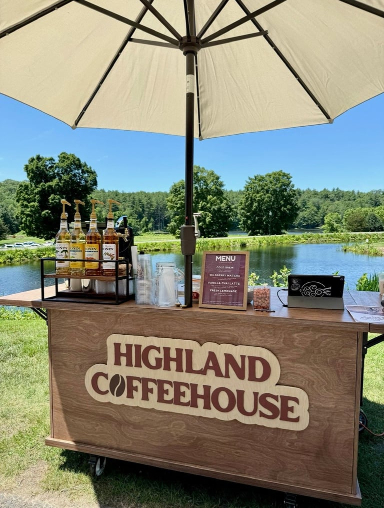 Highland Coffeehouse outdoor pop-up stand with cream umbrella, overlooking a scenic lakeside with trees and green grass